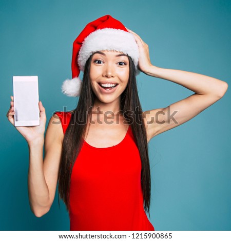 Brunette surprised and excited woman in santa hat is holding smartphone and posing on blue background