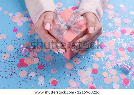 Woman hands holding present box with silver bow on pastel blue background with glitter and confetti. Flat lay style.