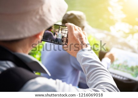 old man travel with backpack in park and take a photo of drawing artist, who draw the landmark view in park, by smart phone in park