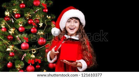 Happy girl in a christmas dress and hat holding the opened present box on christmas tree background