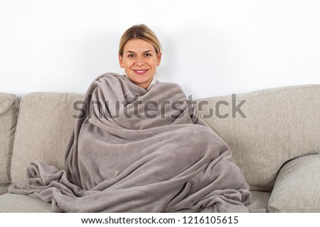 Cute blonde woman relaxing o the sofa wrapped in a soft grey blanket, white wall in the background Royalty-Free Stock Photo #1216105615