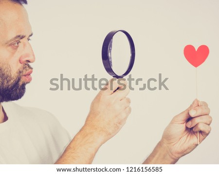 Uncertain man looking at love little heart shape on stick using magnifying glass thinking about romance. Studio shot