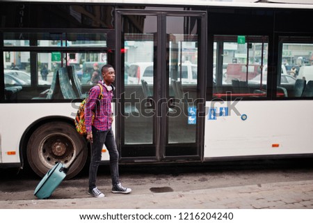 African american man in checkered shirt, with suitcase and backpack. Black man traveler against bus.
