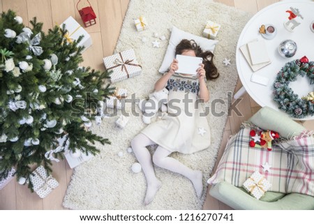 Little girl is lying under the Christmas tree with a tablet on the carpet. View from above