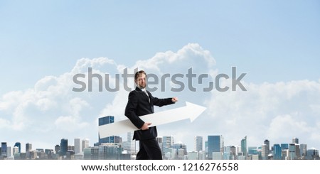 Cropped image of successful young businessman holding big white arrow and pointing away while standing against cityscape view on background