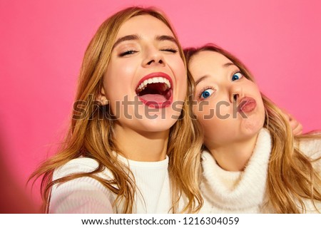 Portrait of two young stylish smiling blond women. Girls dressed in summer hipster clothes. Positive models making selfie on smartphone on pink background