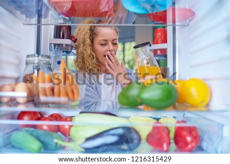 Young Caucasian woman yawning and taking juice from fridge. Morning routine. Picture taken from the inside of fridge.