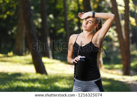 Sportish long-haired young woman wearing black shirt resting before the jogging and drinking water in the sunny park, healthy lifestyle and people concept
