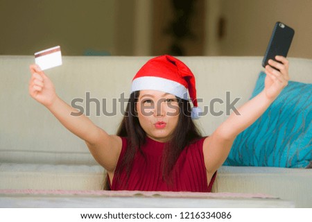 young happy and beautiful Asian Chinese woman in Santa Claus hat holding credit card using mobile phone for Christmas online shopping from home couch smiling cheerful in internet business concept