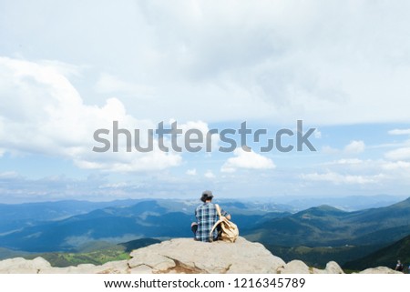 The guy sits back on the edge of the mountain. A man in a plaid shirt sits with his back overlooking the mountains. Beautiful boy in the mountains. A man on the background of a magnificent mountain. Royalty-Free Stock Photo #1216345789