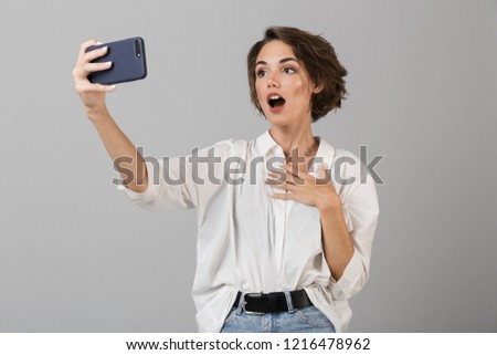 Photo of a shocked young woman posing isolated over grey background make selfie by mobile phone.