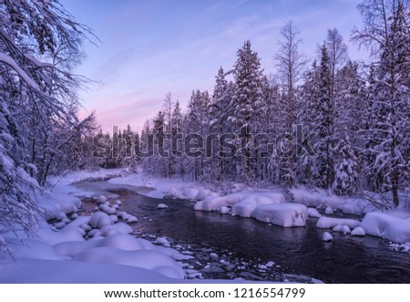 Stunning winter Lapland landscape with beautiful sunset, non-frozen river and snow-covered forest. Untouched white snow at riverbanks. Fantastic ice and frost natural decor on branches over water.