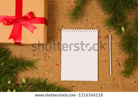 A gift with a red bow, next to a fir branch with cones and a notepad with a pen on a rough embossed background.