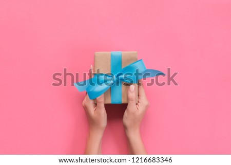 Woman hands give wrapped valentine or other holiday handmade present in paper with blue ribbon. Present box, decoration of gift on pink table, top view with copy space. Royalty-Free Stock Photo #1216683346