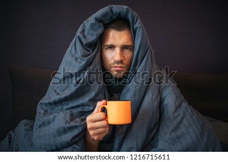 Picture of young man sits on bed and covered up with dark blue blanket. He holds orange cup of tea. Guy looks on camera. He is serious. Young man is emotionless. Royalty-Free Stock Photo #1216715611