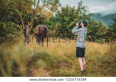 stylish bearded man in a hat and a gray jacket photographing nature among the ouu in the mountains, against the background of a blurred horse and evening slopes of the Carpathian mountains