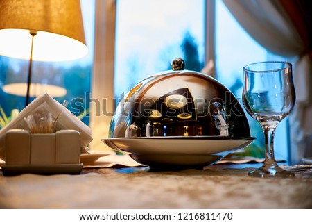 Сloche with dish close-up on a serve table against the background of the window.Deep metal dishes with a lid for serving cooked food on the table. Royalty-Free Stock Photo #1216811470