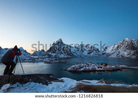 A male photographer uses camera and tripod to capture a beautiful landscape picture at twilight in Lofoten. Winter  scenery with snow-covered mountains, fjord and the fisherman's cabins at Sakrisøy