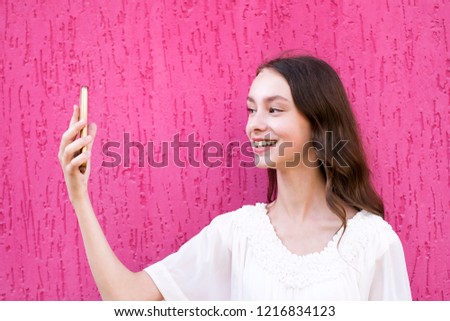 Portrait of pretty cheerful brunette model taking selfie on smartphone standing on bright pink background. Smiling young woman in white outfit. Technology and positivity concept