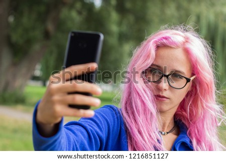 female selfie concept shot of beautiful adult girl portrait with simple makeup, glasses and vivid pink hair and black mobile phone device in her hand in outdoor nature park background 