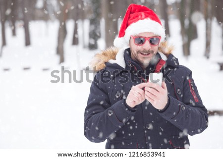 Portrait of young man with santa hat and sunglasses on snow day sending sms