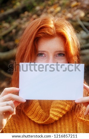 Portrait of beautiful young woman with red hair, ginger, in orange sweater smiling holding a blank blank paper in both hands, copy space, place for your text.