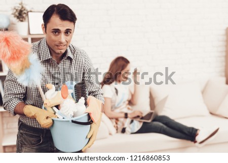 Man and Woman in Apartment. Man Cleans Room. Male with Cleaner Accessories. Male with Dust Brush in Hands and Bucket. Woman Control Cleaning Rrocess. Cleaning Process. Man Cleans and Woman Rests.
