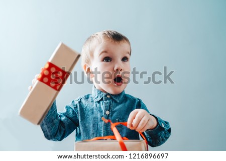 Christmas concept. Cute little baby boy dressed  in demin shirt looking very happy and surprised unboxing holiday presents. Plain grey backround.