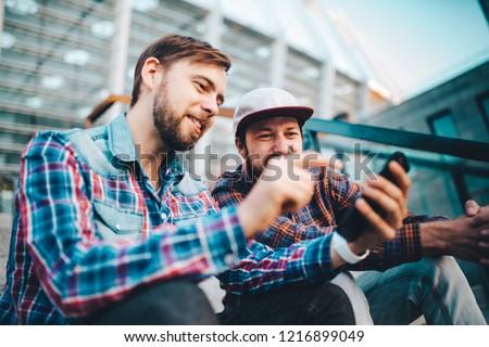Two friends sitting on football stadium steps and making bets using gambling mobile application. Men watching online broadcast waiting for winner results ready to celebrate victory.