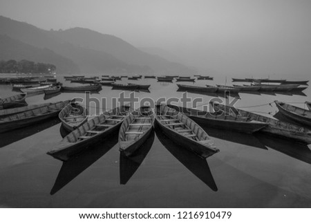 Black and white photo of boats at shore of beautiful Phewa lake with misty landscape on background. Pokhara, Nepal