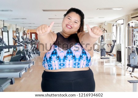 Picture of happy fat woman wearing sportswear while giving thumbs up at the camera and standing in the fitness center