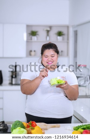 Picture of obese woman looks happy while eating a bowl of healthy salad in the kitchen