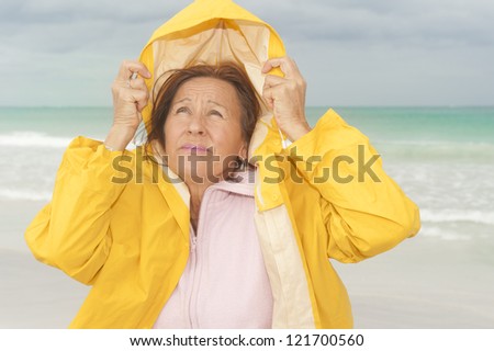 Portrait attractive and worried mature woman with yellow  protective raincoat, isolated with ocean and cloudy autumn sky as blurred background and copy space.