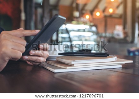 Closeup image of woman's hands holding and pointing at smart phone with notebook on wooden table in cafe