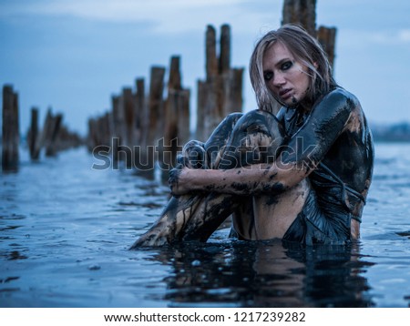 Portrait of Blonde Caucasian Girl Sitting Smeared in a Healthy Black Mud in old Firth with Wooden Posts for Salt Production