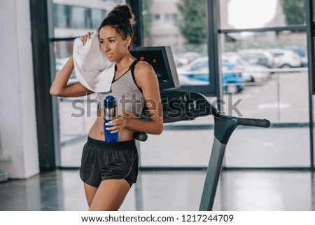 african american sportswoman with sport bottle wiping head by towel near treadmill at gym