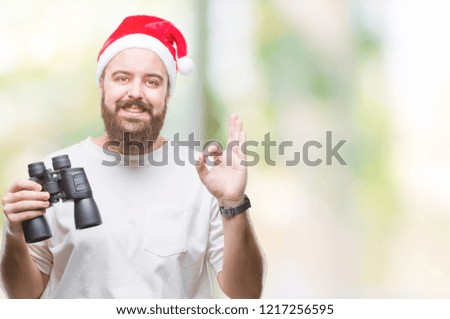 Young caucasian hipster man wearing christmas hat looking though binoculars over isolated background doing ok sign with fingers, excellent symbol