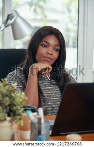 Beautiful black woman watching laptop pensively working at table in modern office