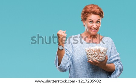 Atrractive senior caucasian redhead woman eating popcorn over isolated background screaming proud and celebrating victory and success very excited, cheering emotion