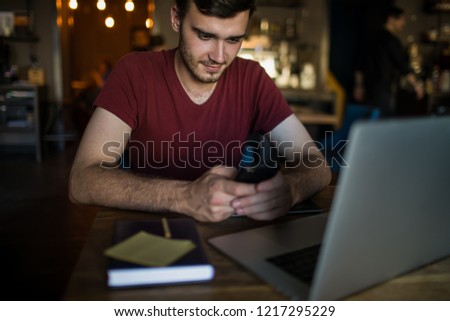 Man owner online business in internet reading notification on mobile phone while sitting with laptop computer in modern restaurant.Male professional marketing coordinator checking e-mail on smartphone