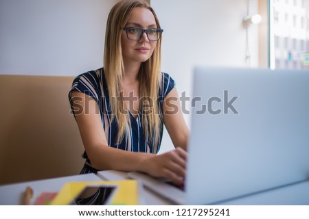Young European woman commercial real estate analyst using applications on laptop computer while sitting in coworking space. Female in glasses experienced freelancer working distance on netbook  
