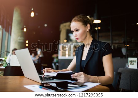 Successful woman jurist using textbook while reading article on web site via laptop computer, sitting in modern restaurant. Confident female marketing specialist checking e-mail via portable netbook 