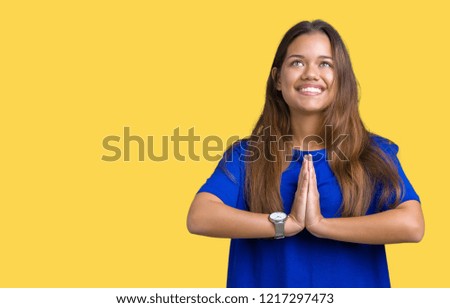 Young beautiful brunette woman wearing blue t-shirt over isolated background praying with hands together asking for forgiveness smiling confident.