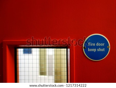 Closeup small warning sign, Fire door keep shut. Selective focus on blue metal badge sign on right side of frame. Space to add text on red surface, blurry mesh grass & white door in backgound. 