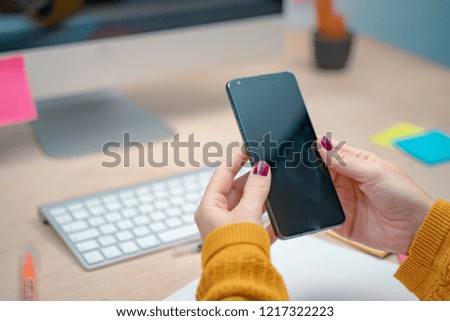 Young female hands holding a black screen smartphone on her workplace at the office. Morning at a creative studio. Graphic design, mock up screens. glasses, pens, keyboard, notebook, colors, modern.