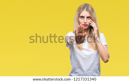 Young beautiful blonde woman talking using smartphone over isolated background with open hand doing stop sign with serious and confident expression, defense gesture