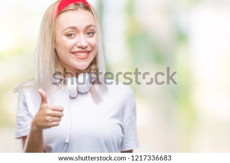 Young blonde woman wearing headphones listening to music over isolated background doing happy thumbs up gesture with hand. Approving expression looking at the camera with showing success.
