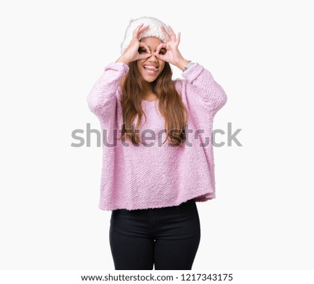 Young beautiful brunette woman wearing sweater and winter hat over isolated background doing ok gesture like binoculars sticking tongue out, eyes looking through fingers. Crazy expression.