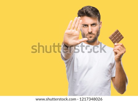 Young handsome man eating chocolate bar over isolated background with open hand doing stop sign with serious and confident expression, defense gesture