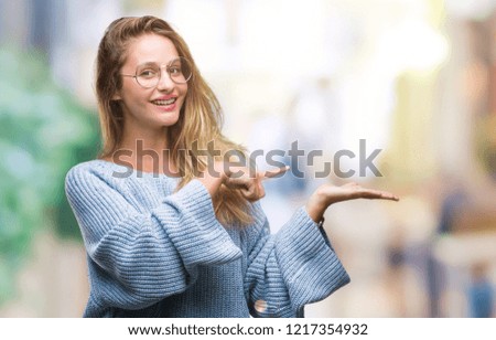 Young beautiful blonde woman wearing sweater and glasses over isolated background amazed and smiling to the camera while presenting with hand and pointing with finger.